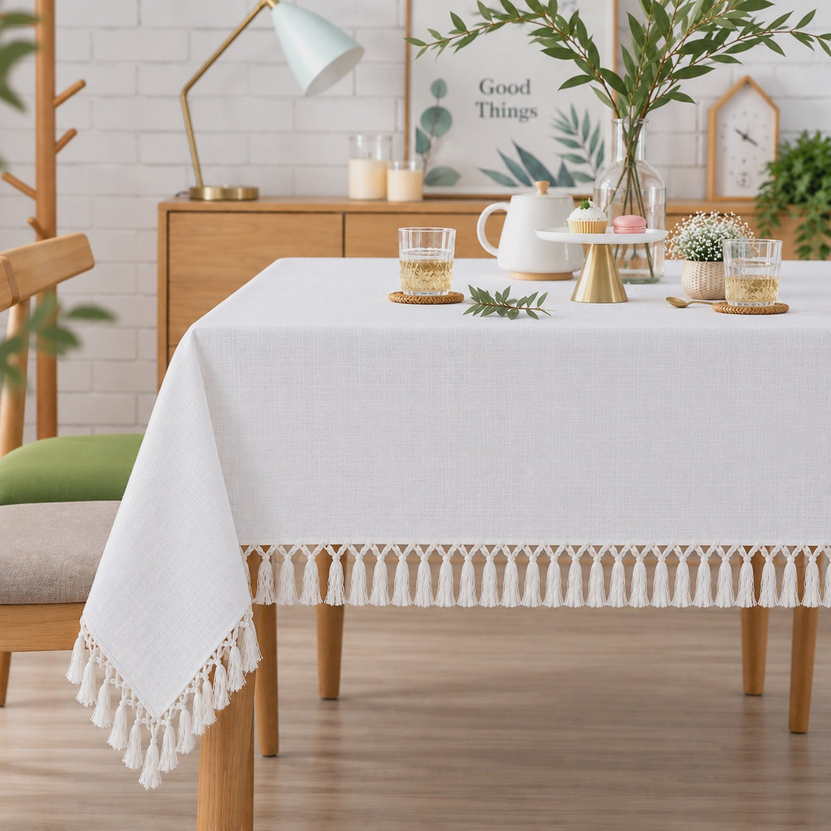 A white tablecloth with a decorative tassel fringe along the bottom edge, styled on a wooden dining table in a bright, modern room.