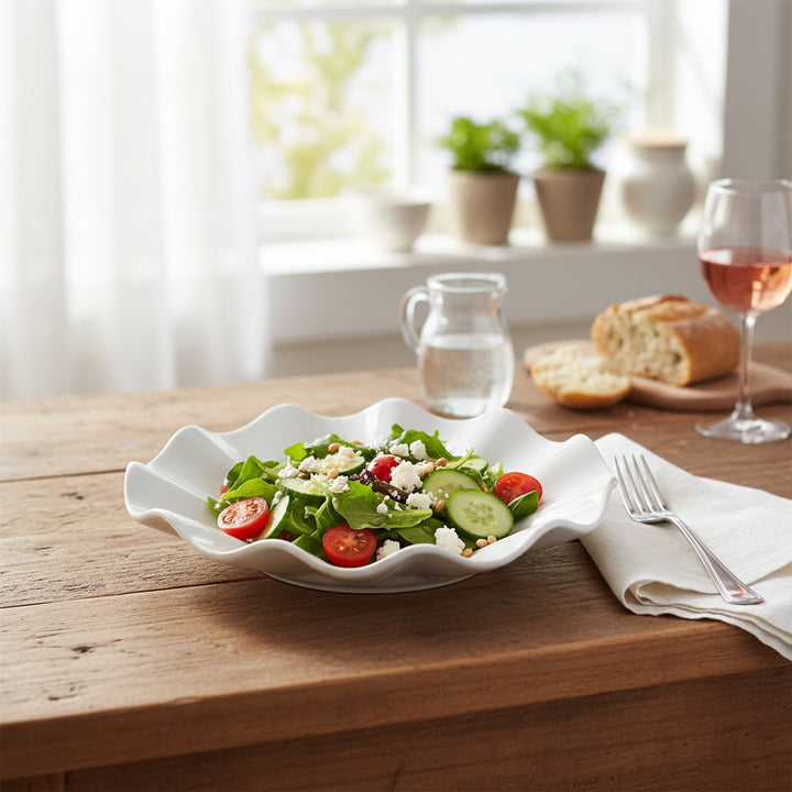 White scalloped-edge bowl with a salad on a wooden table, accompanied by a glass of wine and bread.