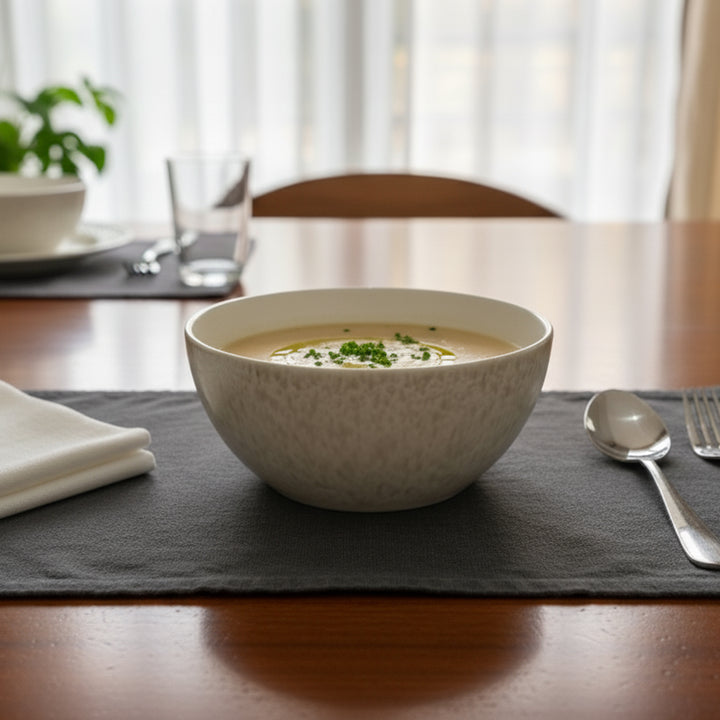 Bowl of soup on a table with a placemat, spoon, and fork.