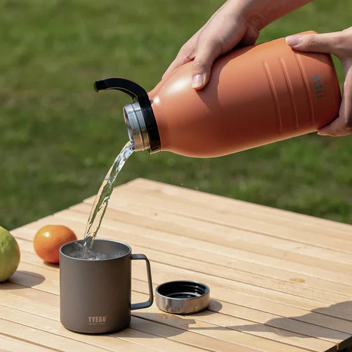 Person pouring water from a terracotta-colored bottle into a gray mug on a wooden table with a green outdoor background.
