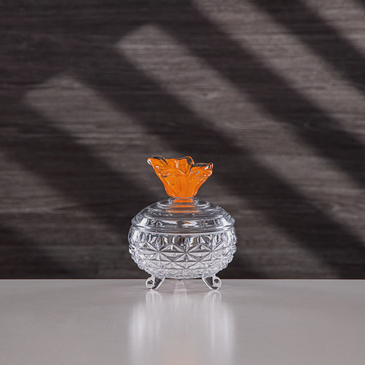 Decorative glass container with an orange top on a reflective surface with a dark textured background