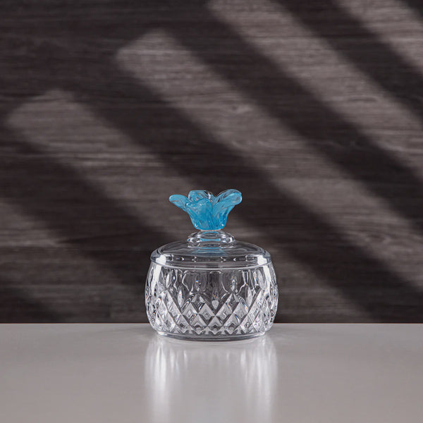 Crystal jar with a blue decorative lid on a reflective surface with a dark, patterned background
