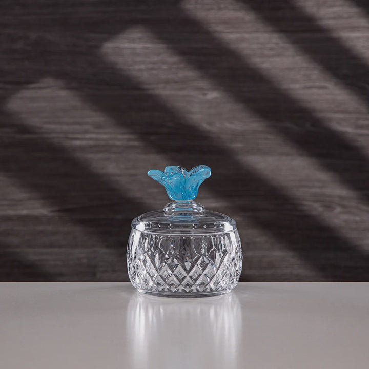 Crystal jar with a blue decorative lid on a reflective surface with a dark, patterned background