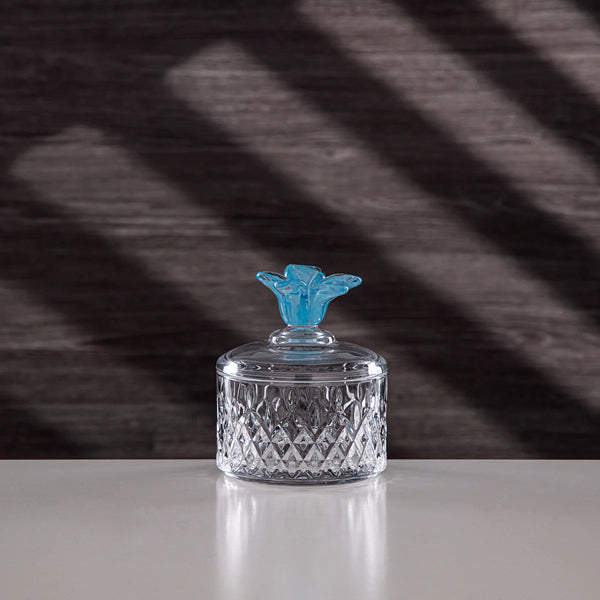 Clear glass container with a blue decorative top on a reflective surface with a dark, patterned background