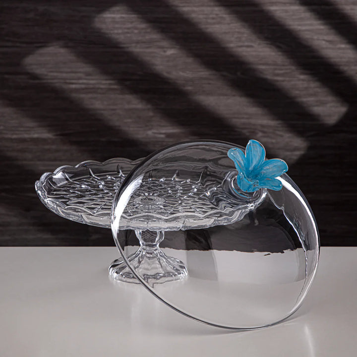Clear glass cake stand with a blue flower decoration on a black and white striped background