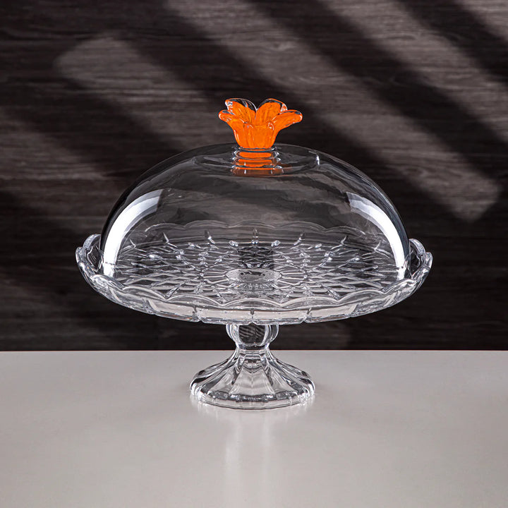Clear glass cake stand with an orange floral finial on a dark background