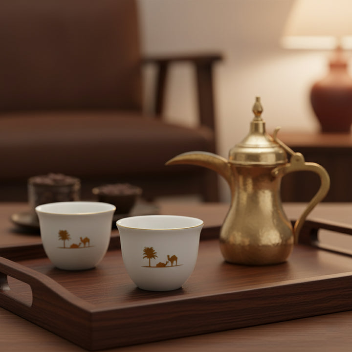 Two white cups with camel design and a gold teapot on a wooden tray in a warm-toned room.