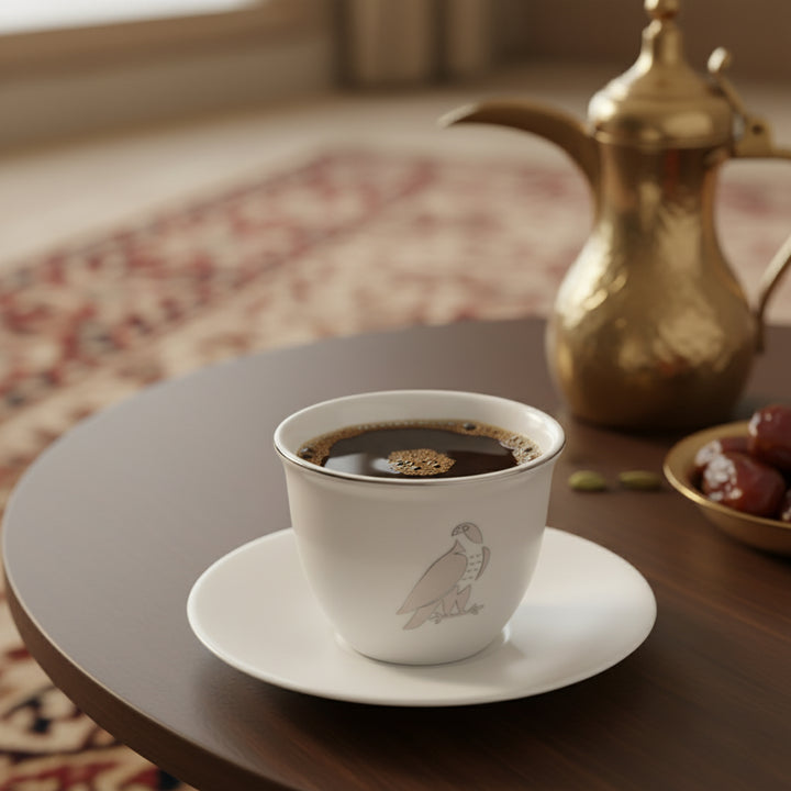 White coffee cup with a bird design on a saucer, placed on a wooden table with a gold teapot and dates in the background.