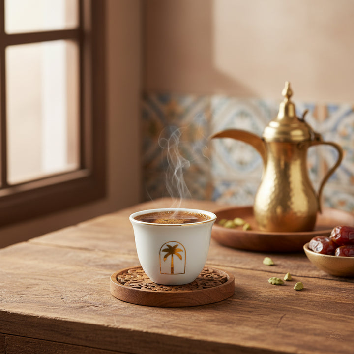 Steaming cup of coffee with a palm tree design on a wooden table, accompanied by a gold teapot and dates.