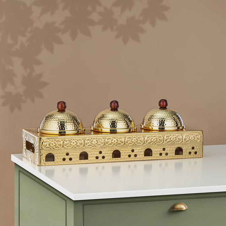 Decorative gold tray with three domed covers on a white surface against a beige wall.