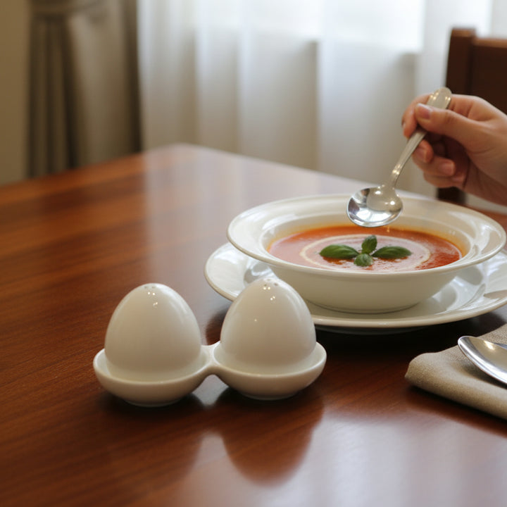 Tomato soup with cream and basil leaves on a plate, accompanied by an egg holder and a spoon on a wooden table.