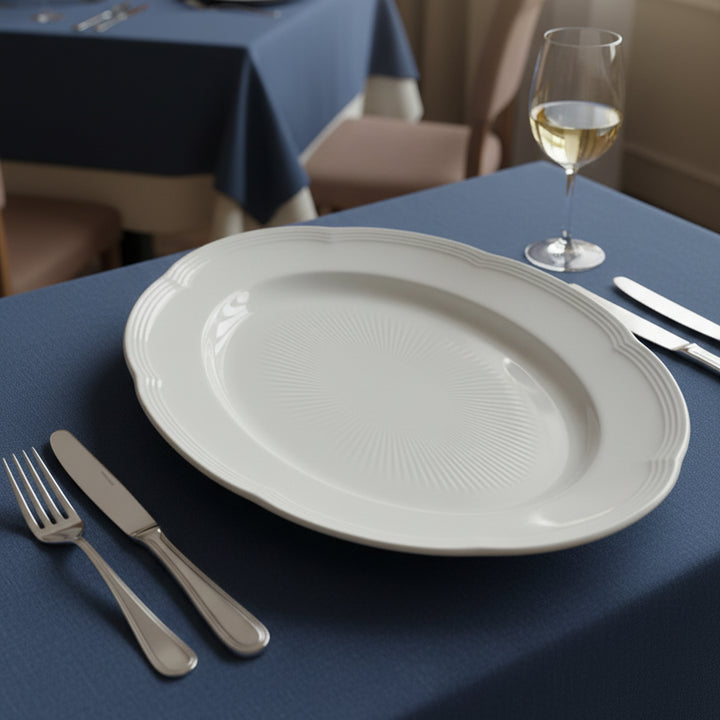 White plate on a blue tablecloth with silverware and a glass of white wine.