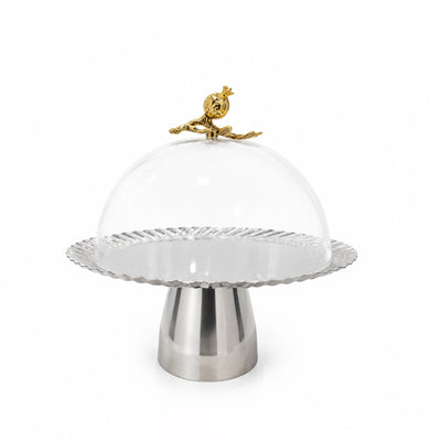 A modern pedestal cake stand featuring a fluted silver metal base, a clear glass dome lid, and a decorative gold metal bird finial on top, set against a plain light gray background.