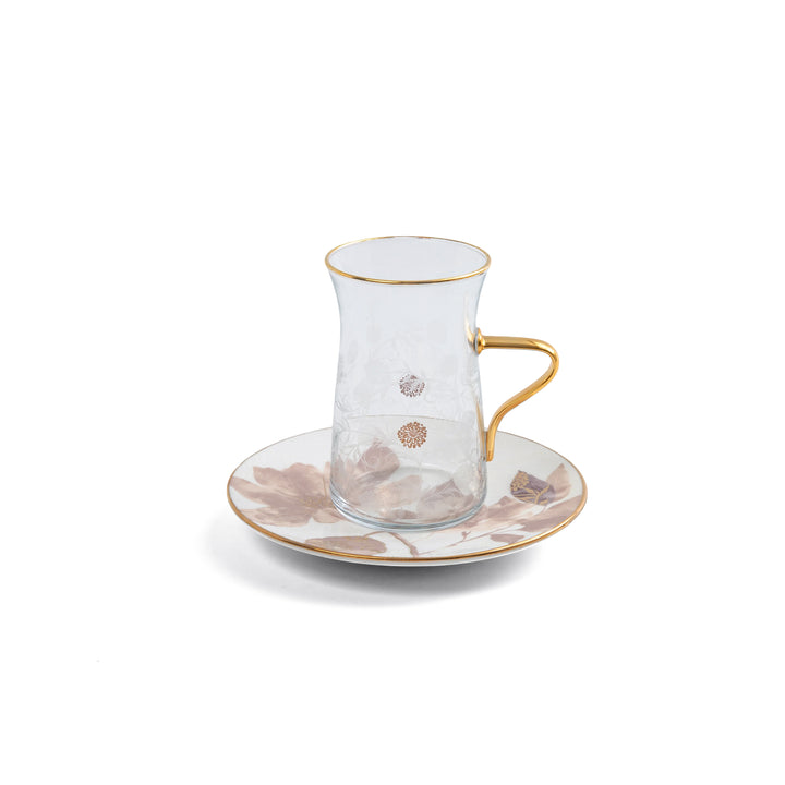 Clear glass teacup with gold rim and handle on a matching saucer with floral design on a white background