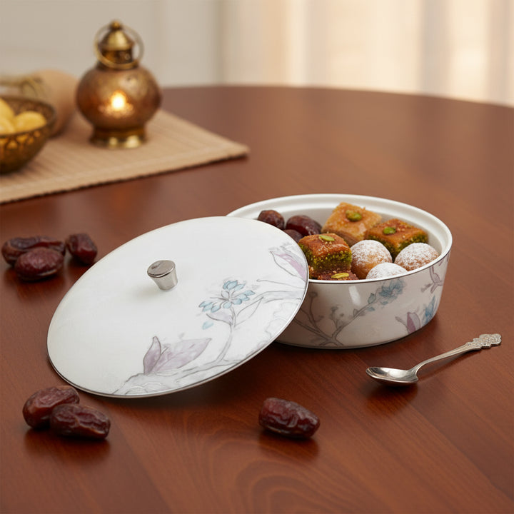 Decorative bowl with lid containing pastries on a wooden table with dates and a spoon.