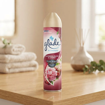 A can of Glade Air Freshener, Blooming Peony & Cherry, standing on a light wooden table in a softly lit bathroom or bedroom setting with folded towels and a reed diffuser in the background.
