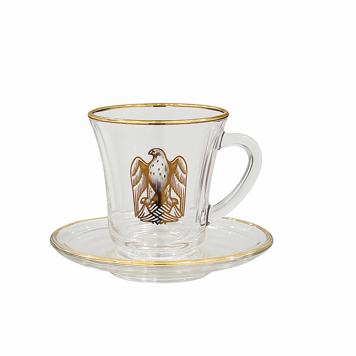 Clear glass cup with gold rim and handle, featuring a gold emblem on a white background