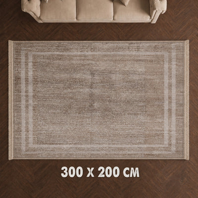 A rectangular area rug in a warm taupe-beige tone with a subtle textured weave and a faded geometric border, shown from above on a dark wood floor with a sofa partially visible at the top and size text '300 X 200 CM' at the bottom.