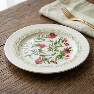 A round white ceramic plate with a hand-painted pomegranate and leaf pattern, styled on a rustic wooden table next to a gold fork and a folded beige napkin.