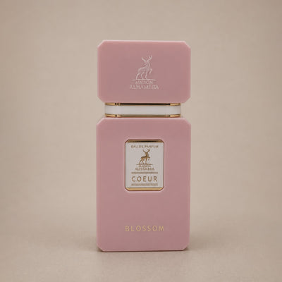 A pink rectangular perfume bottle with a matching cap, featuring gold lettering and a small white label on the front, photographed against a plain beige background.