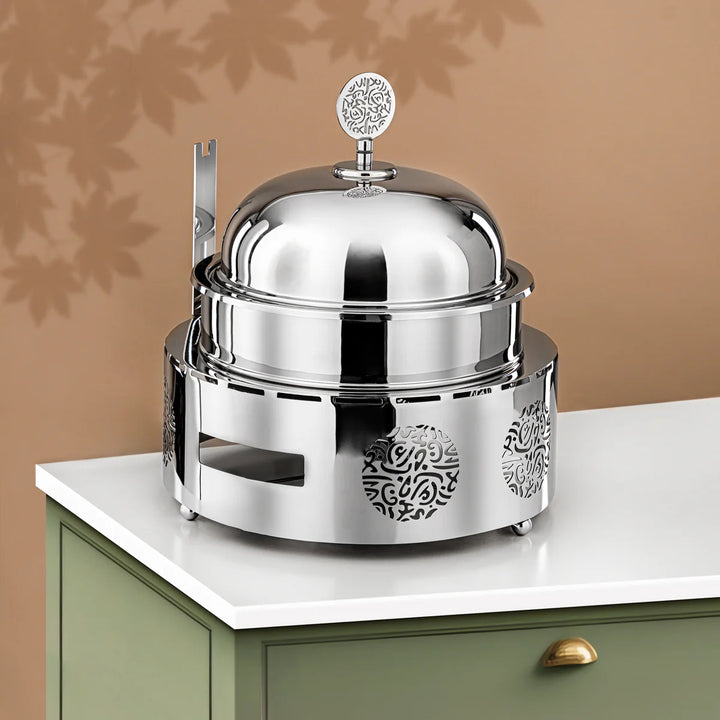 Silver chafing dish with decorative patterns on a white surface against a beige wall.
