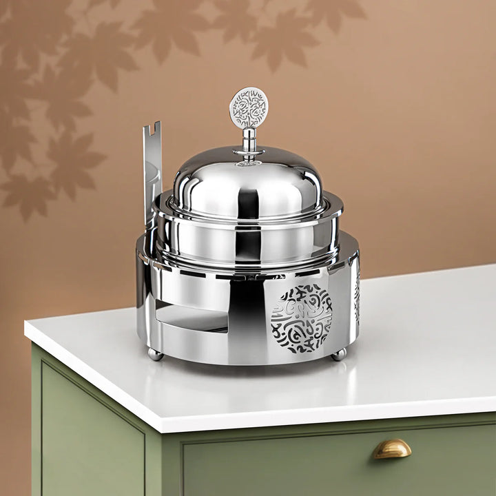 Silver chafing dish with decorative elements on a white surface against a beige wall.