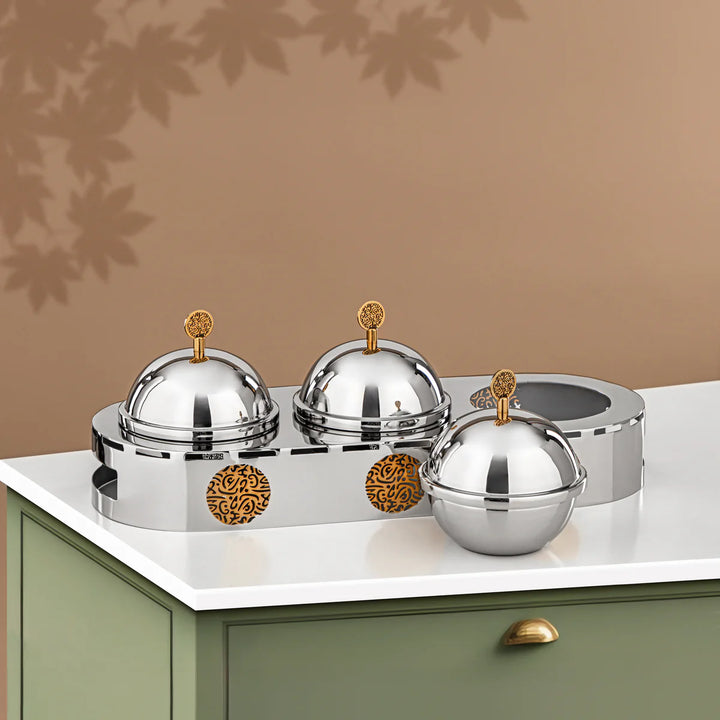 Set of silver domed containers with gold accents on a white surface against a brown wall.