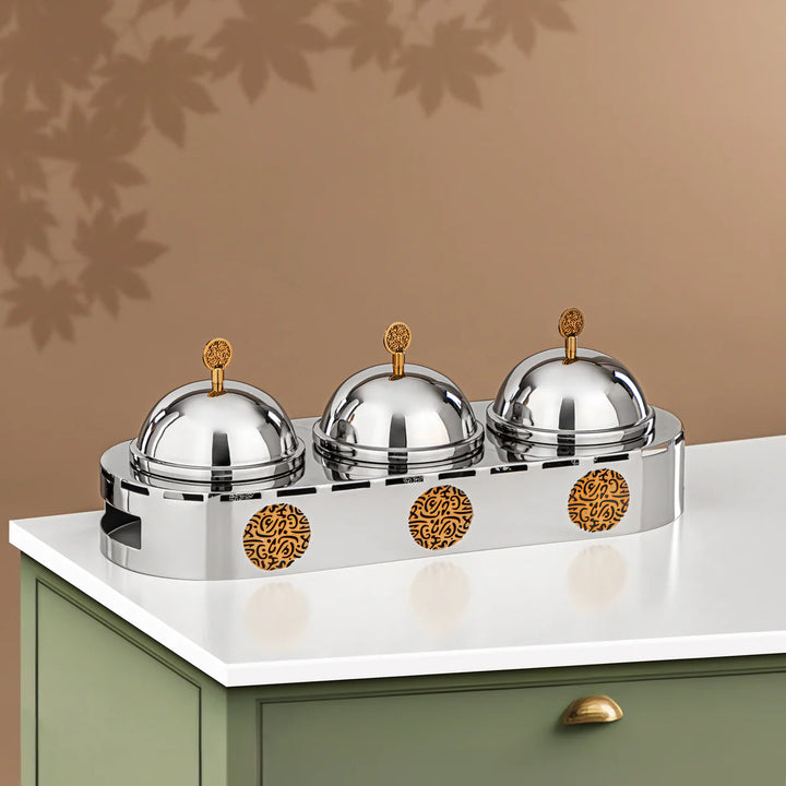 Three silver food covers with gold domes on a decorative stand against a brown wall.