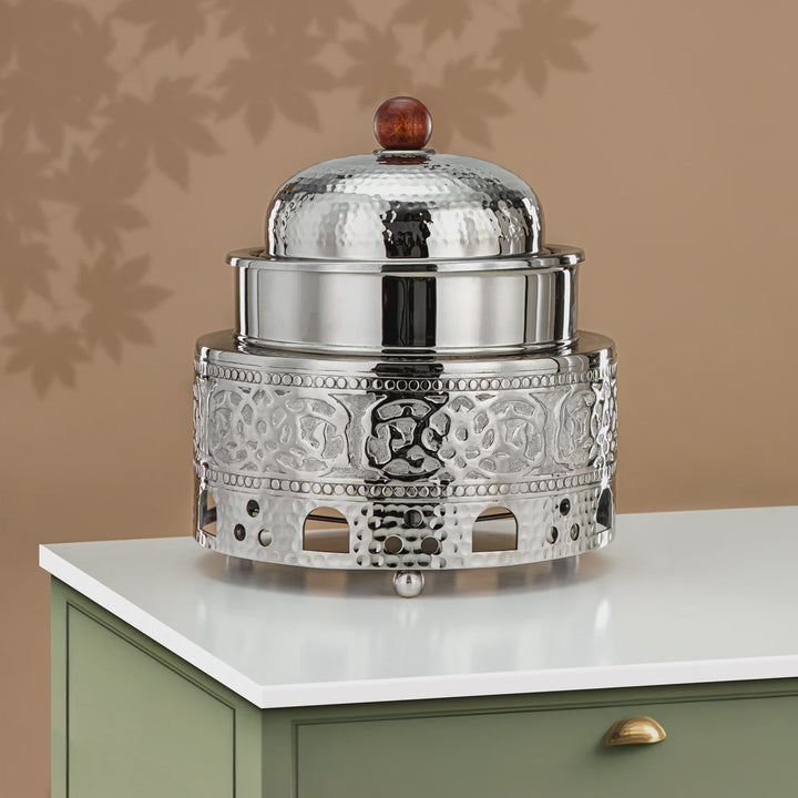 Decorative silver container with a glass lid on a white surface against a beige wall.