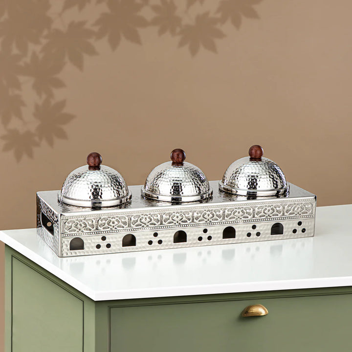 Decorative silver tray with three domed covers on a green cabinet.