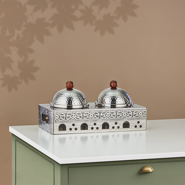 Decorative silver box with two domed lids on a white surface against a beige wall.