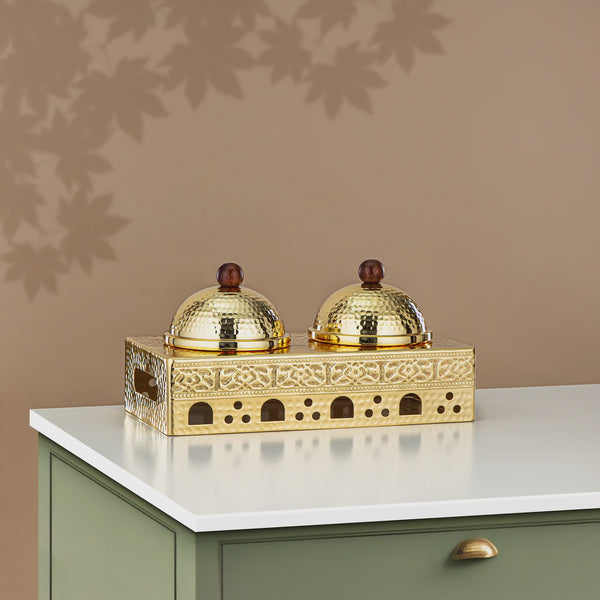 Decorative gold holder with two domed containers on a white surface against a brown wall.
