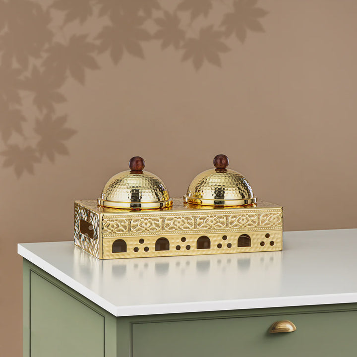 Decorative gold holder with two domed containers on a white surface against a brown wall.