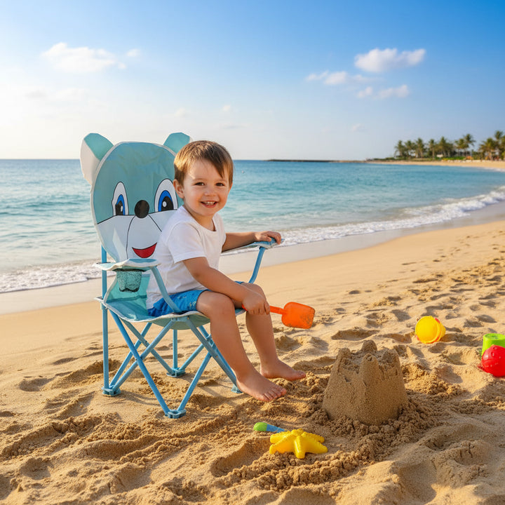 Child sitting on a blue cartoon chair at the beach with sand toys and a sandcastle.