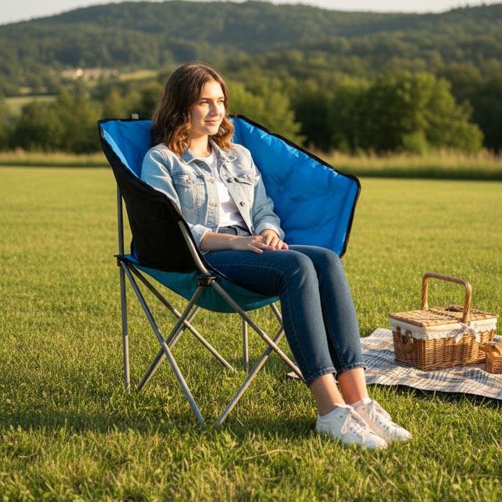 Woman sitting in a blue camping chair in a grassy field with a picnic basket.