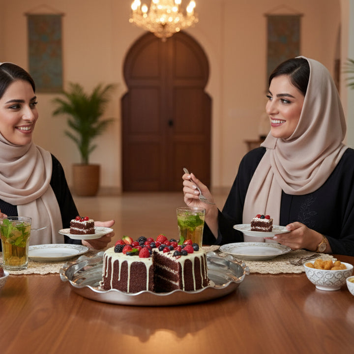 Two women sitting at a table with a large chocolate cake and drinks in a decorated room.