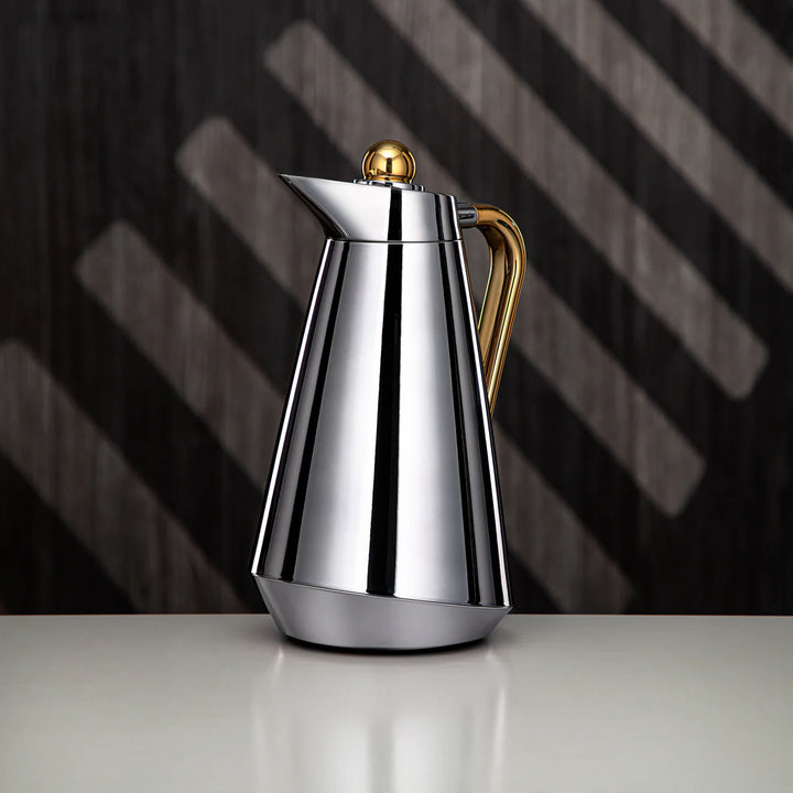 Silver coffee pot with gold accents on a reflective surface with a dark, striped background