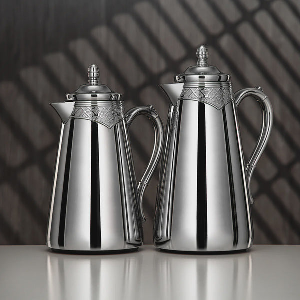 Two silver coffee pots on a reflective surface with a dark, patterned background