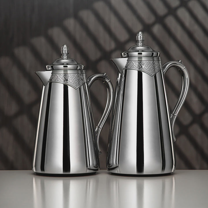 Two silver coffee pots on a reflective surface with a dark, patterned background