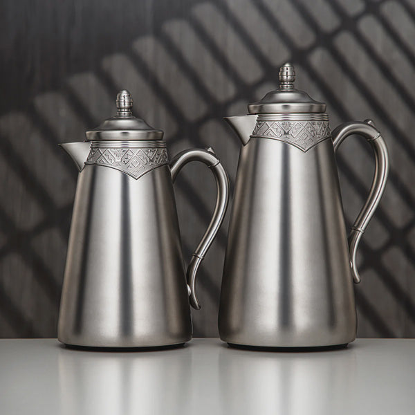 Two silver coffee pots with decorative lids on a reflective surface.