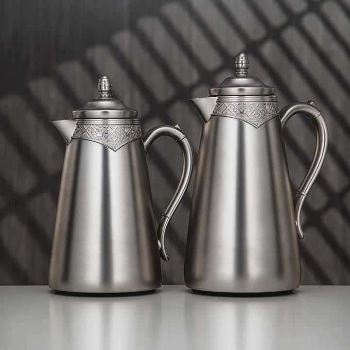Two silver coffee pots with decorative lids on a reflective surface.