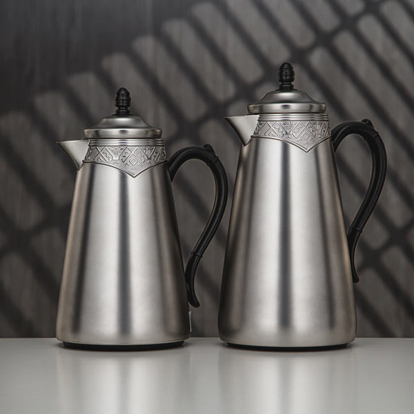 Two silver coffee pots with black handles on a reflective surface with a dark, patterned background.