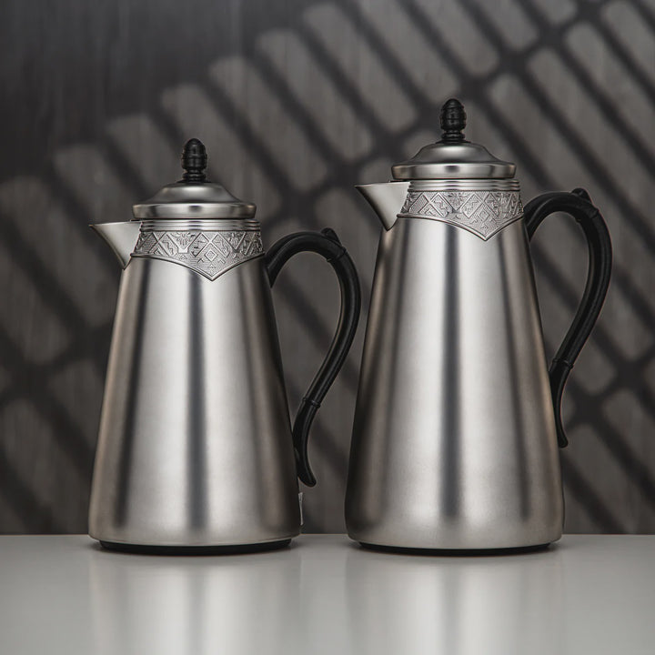 Two silver coffee pots with black handles on a reflective surface with a dark, patterned background.