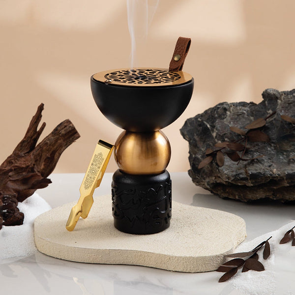Decorative incense burner with gold and black design on a marble surface with rocks and leaves.